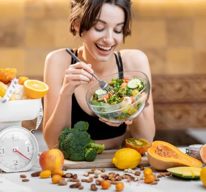 A smiling woman eating a bowl of salad beside fresh fruit and vegetables